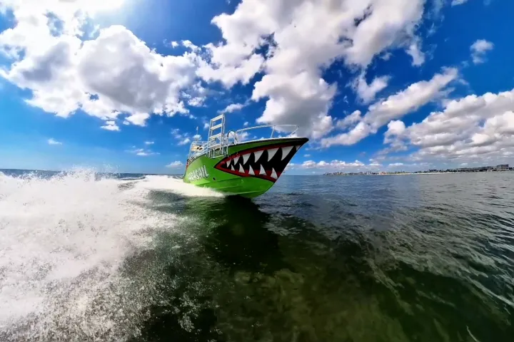 Green boat with shark mouth design speeding on water under blue sky.