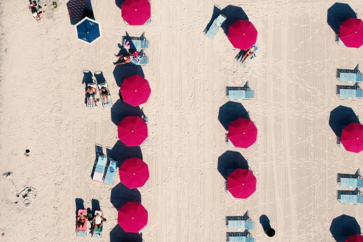 Aerial view of beach with red umbrellas, chairs, and sunbathers on sand.